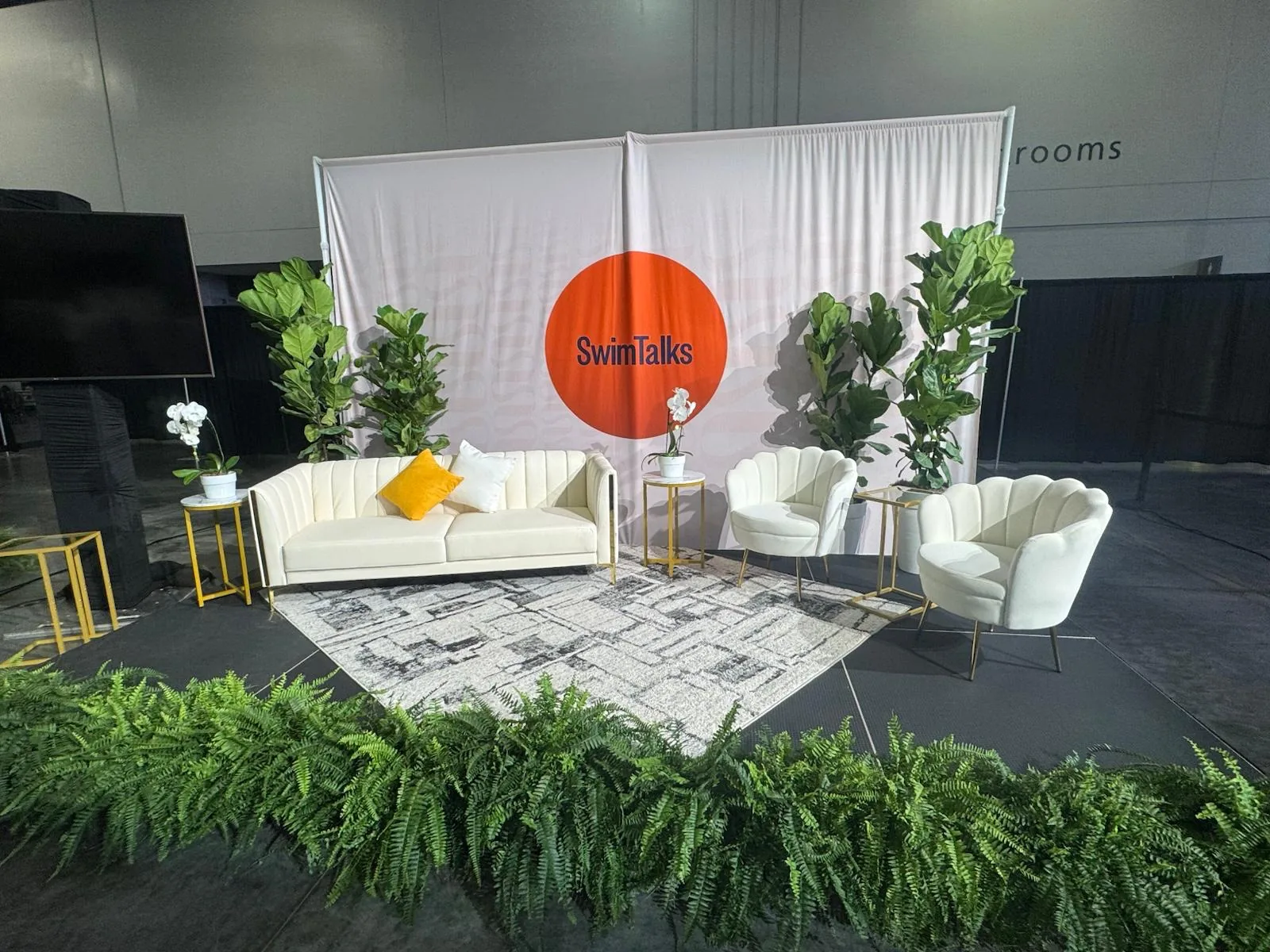 White lounge seating framed by fiddle-leaf figs and a run of Boston ferns at a trade show booth